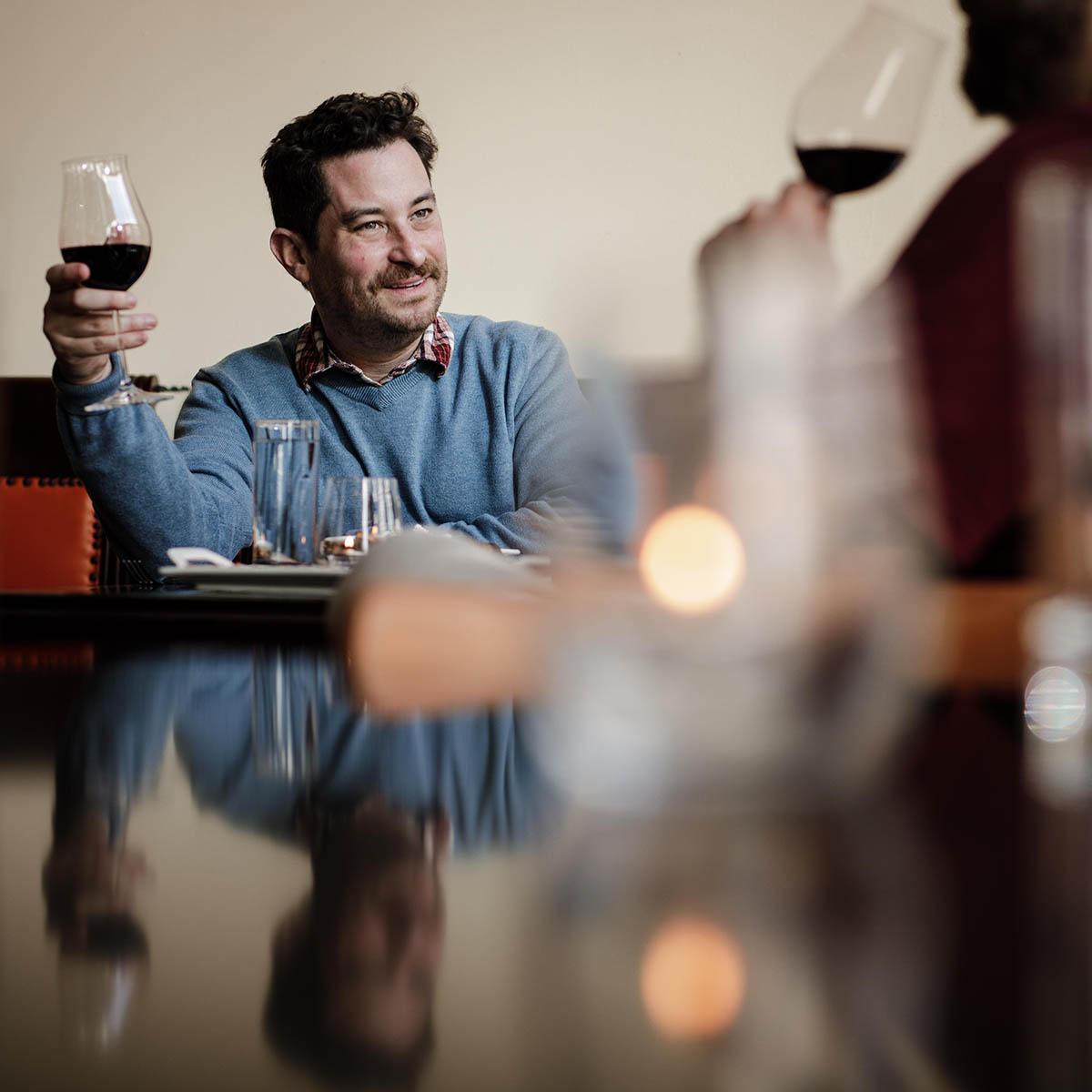 Photo of two men holding glasses of red wine, looking at one another in conversation across a table