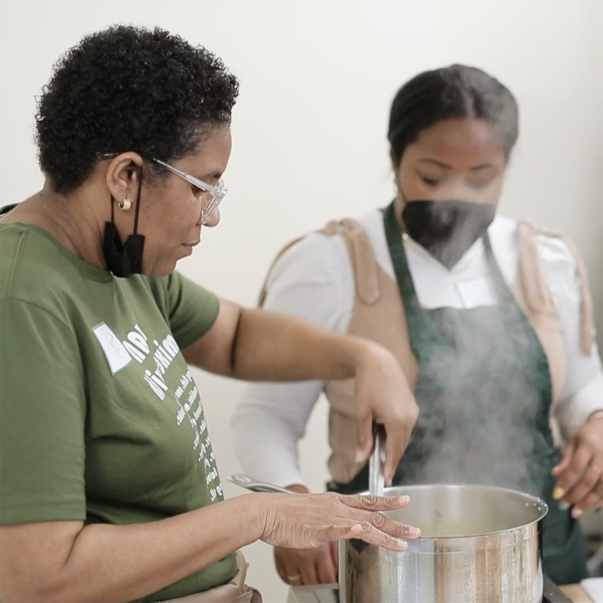 Photo of a Black woman, Toni Simpson, stirring food in a pot and demonstrating to a younger Black woman in an apron and mask who is observing